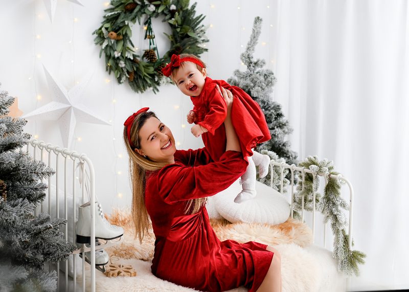 Mama w czerwonej sukni podrzuca córeczkę w czerwonej sukience na tle białych zasłon i choinek – jasna świąteczna scenka w studiu pod Tarnowem